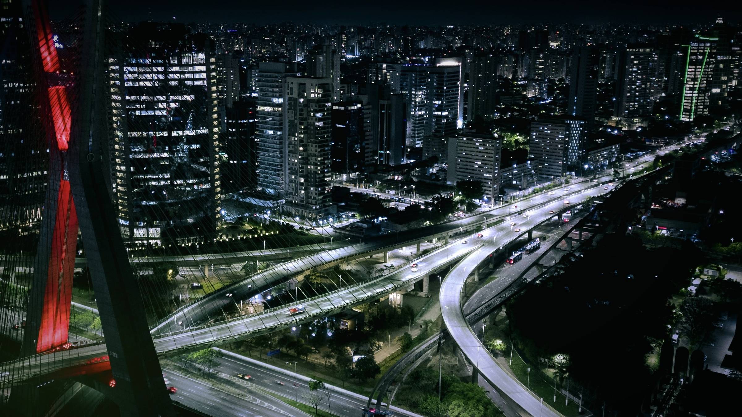 Vista aérea noturna de uma metrópole brasileira, com viadutos iluminados e uma ponte estaiada